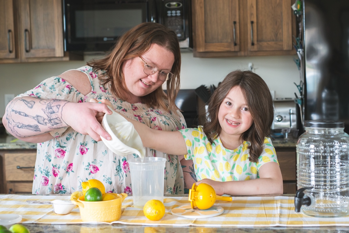 daughter smiles at camera while helping mom pour fresh lemonade