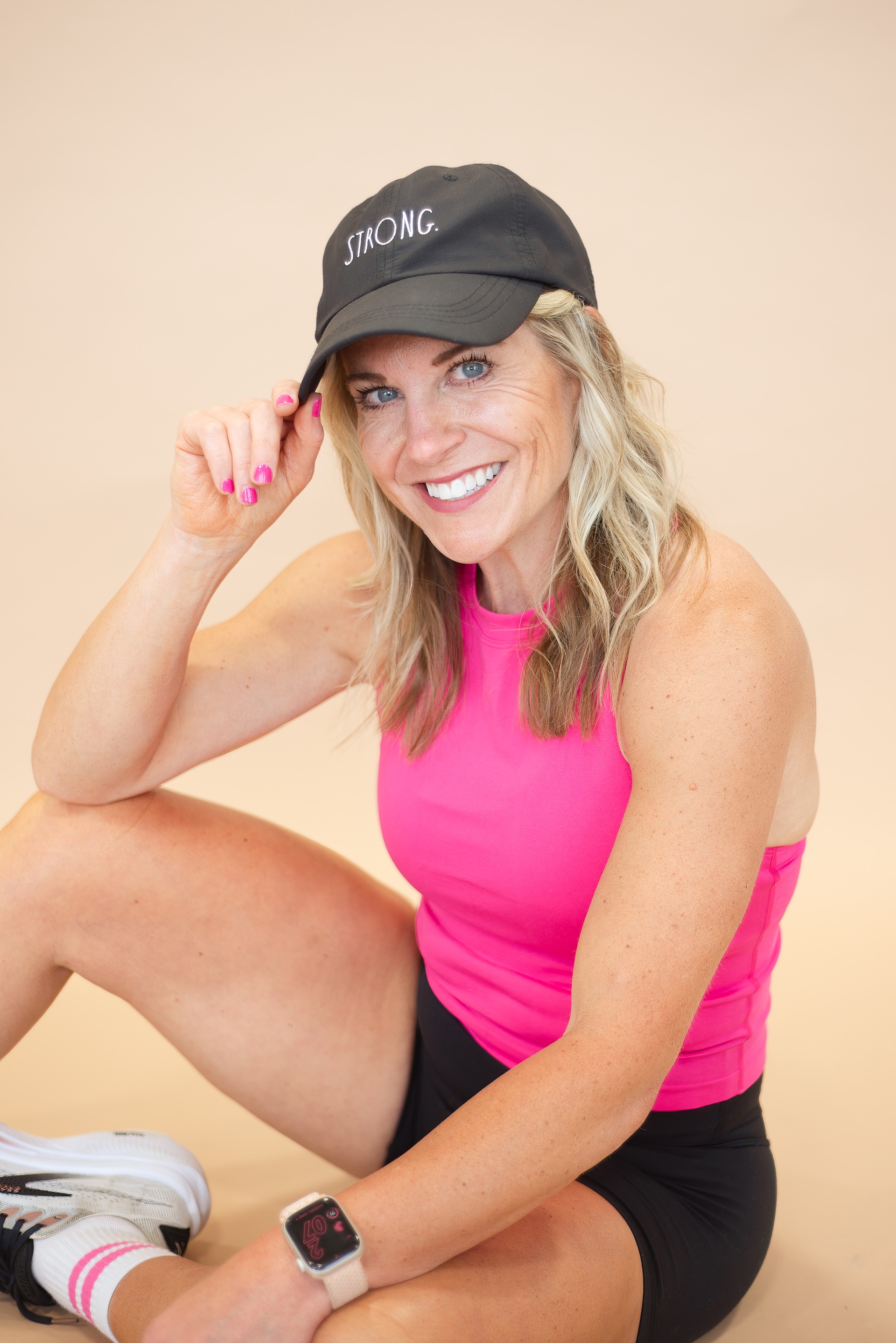 Inspire Health and Wellness wearing a black baseball cap and a pink tank top, smiling as she adjusts the brim of her hat.